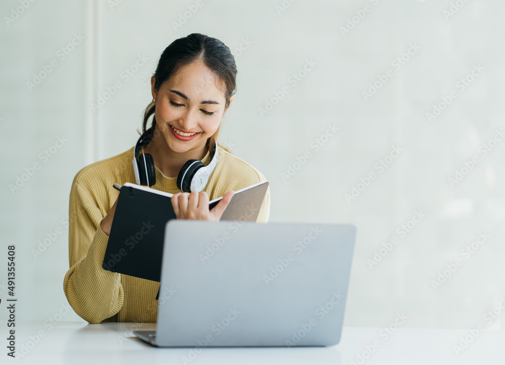 微笑的亚洲年轻女性戴着耳机看着笔记本电脑屏幕听并学习在线课程
