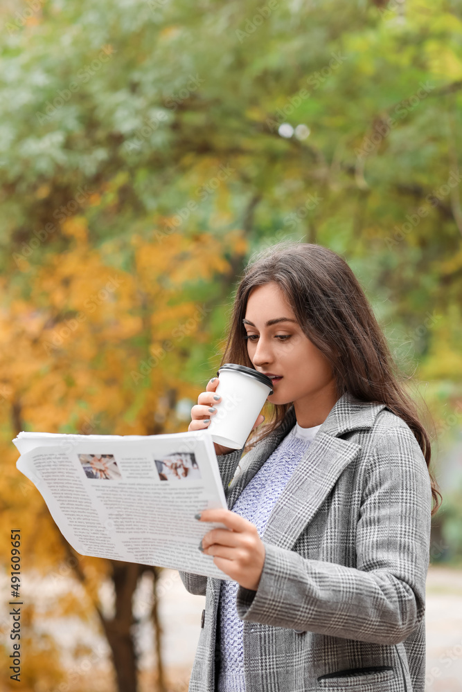 美丽的年轻女子在秋季公园喝咖啡读报