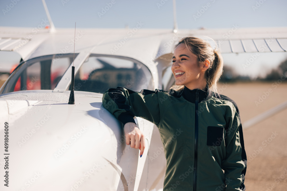 Delighted caucasian female flying officer, looking to the side, leaning on the plane.