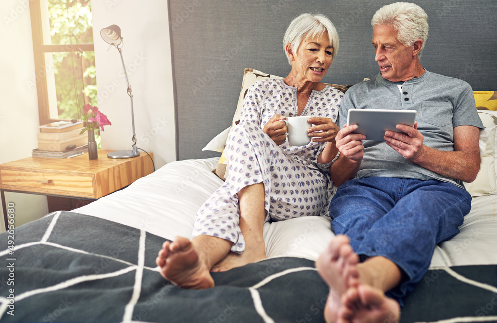 让我们在网上预订假期。一对老年夫妇躺在床上使用平板电脑的照片。