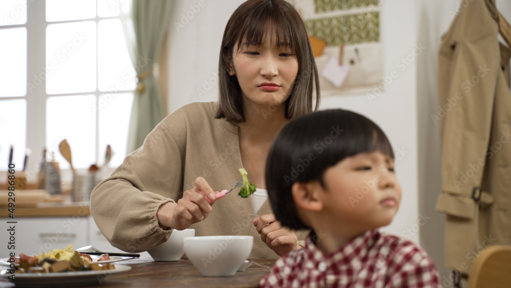 沮丧的亚洲母亲拿着一把叉子放蔬菜，让他的小儿子咬一口迪尼