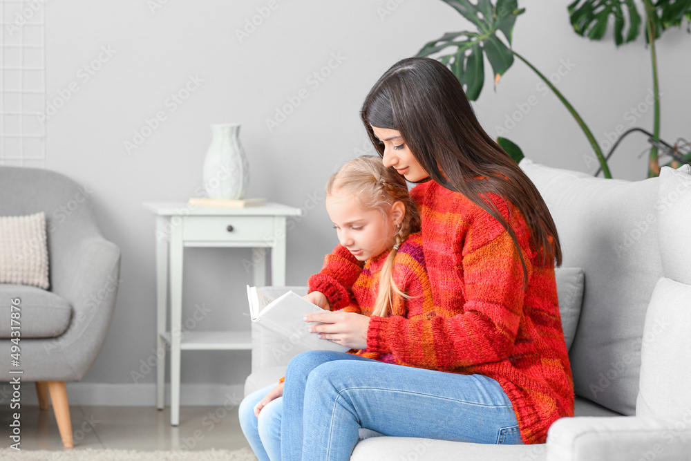 穿着暖和毛衣的小女孩和她的妈妈在家的沙发上看书
