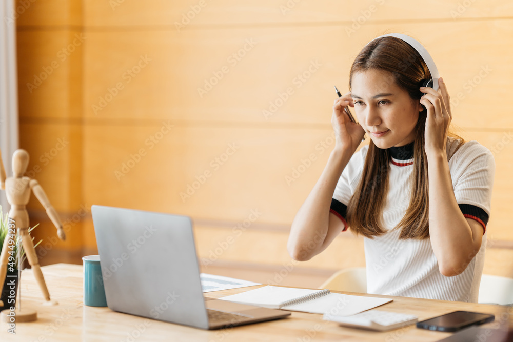 戴着耳机的年轻亚洲女性在家里用笔记本电脑聊天。在家工作