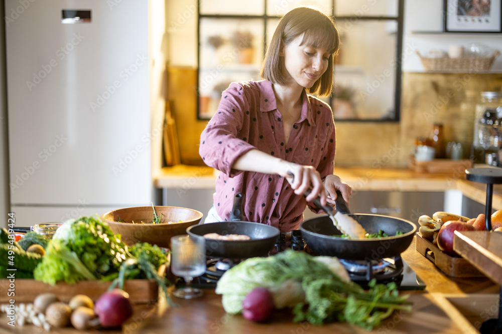 年轻女子在家里的厨房里烹饪健康食品时，在平底锅上炒菜。很多