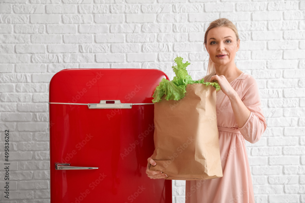 在白色砖墙背景上时尚的红色冰箱附近拿着一袋新鲜产品的女人