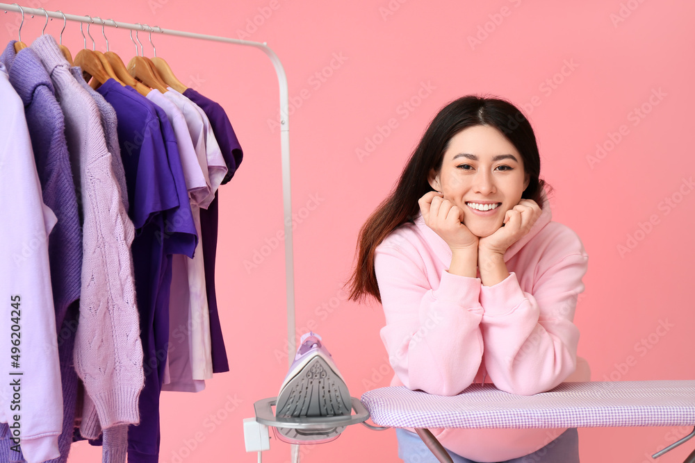 Young Asian woman with rack, clothes and ironing board on pink background