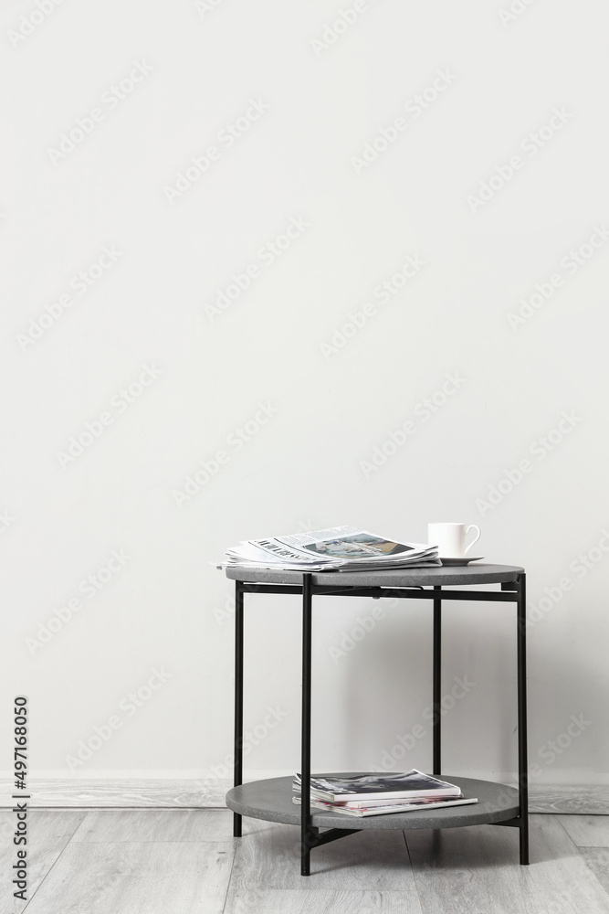 Small table with cup of coffee, newspapers and magazines near light wall