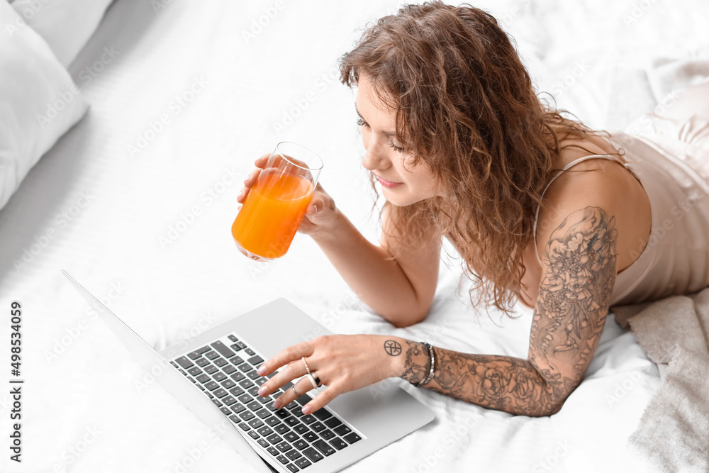漂亮的纹身女人拿着笔记本电脑在卧室里喝果汁