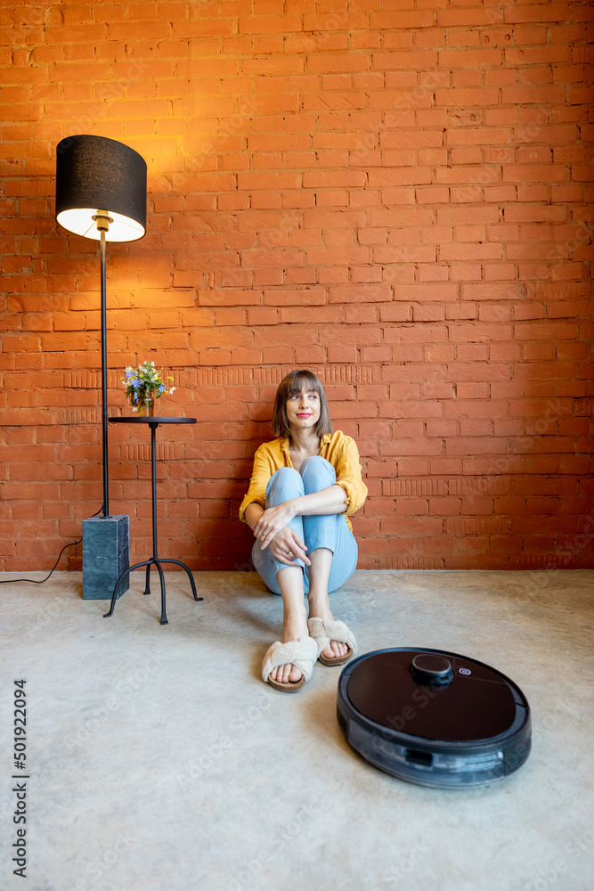 年轻女子坐在地板上放松，机器人吸尘器在家清洁地板。概念