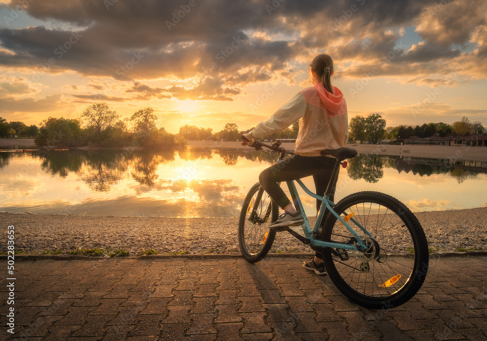 夏天日落时，一名女子在湖边骑山地自行车。五颜六色的风景和运动女孩的骑行