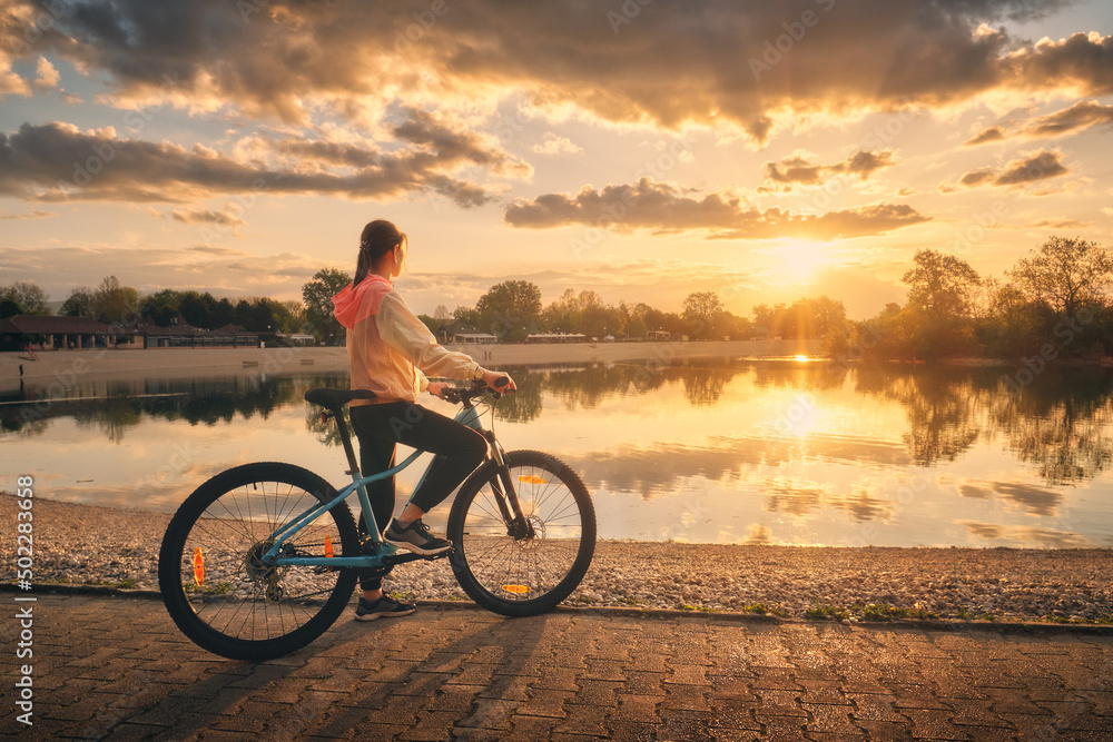 夏天日落时分，在湖边骑山地自行车的女人。色彩斑斓的风景配上运动的女孩ridi