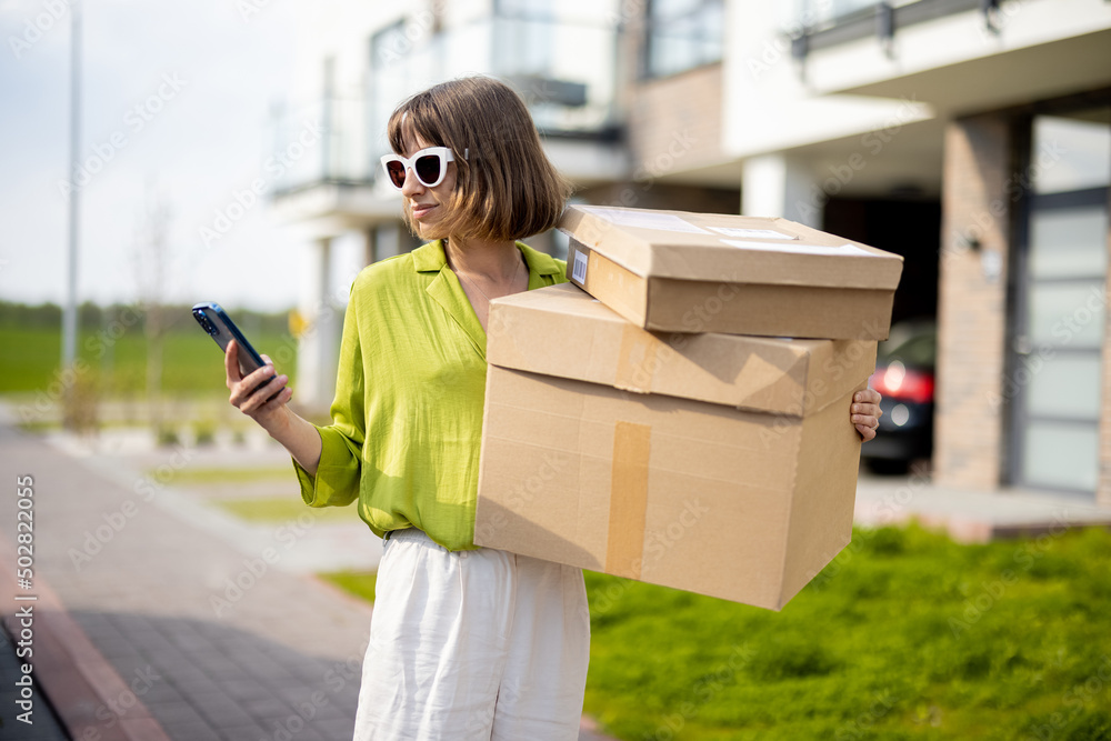 Young stylish woman using phone while standing with parcels on a street outdoors, carrying goods pur