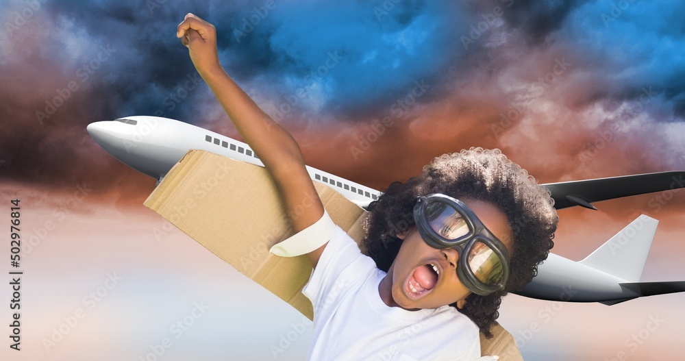 African american boy with cardboard wings and goggles flying and airplane flying in cloudy sky