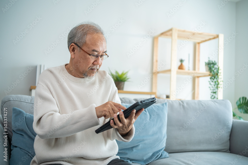 亚洲老年男性在家中客厅使用数字平板电脑。
