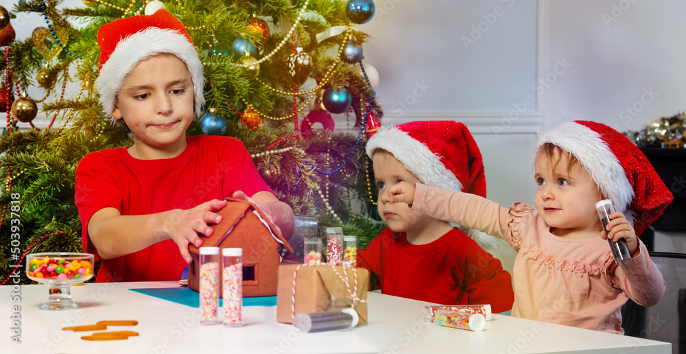 三个孩子戴着圣诞老人帽一起装饰姜饼屋