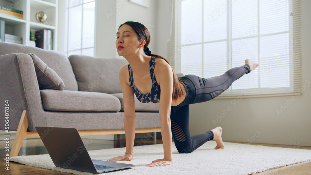 年轻健康的亚洲女性穿着运动服练习瑜伽。在客厅通过笔记本电脑在线训练