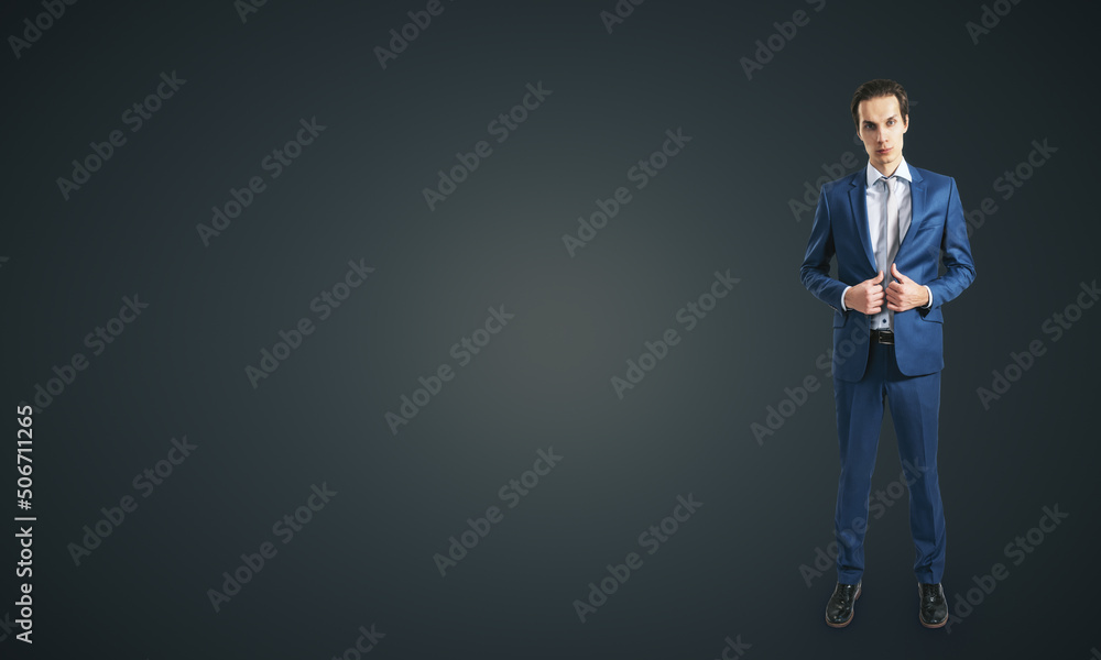 Young self-confident businessman in blue suit and grey tie isolated on dark grey wall background wit