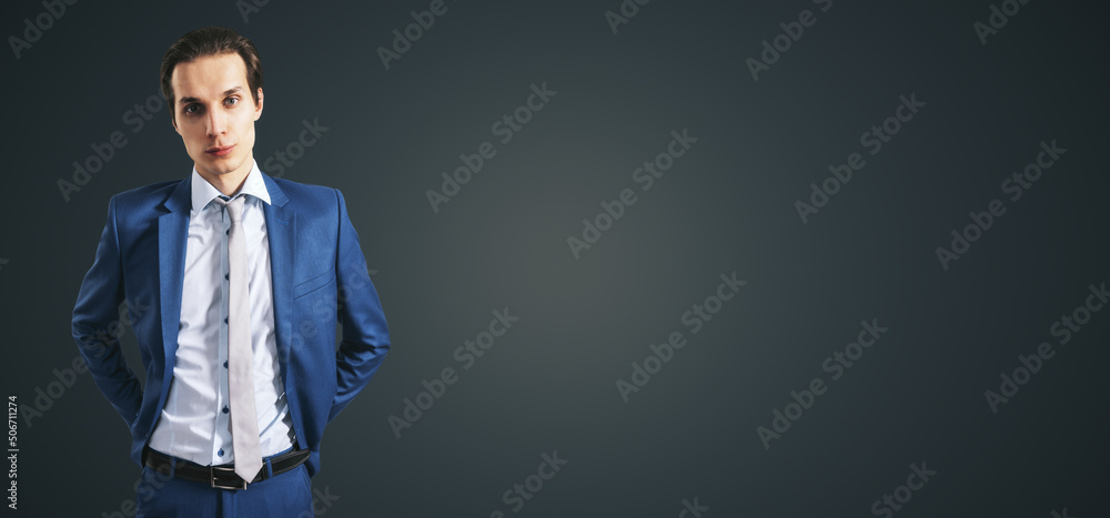 Handsome young businessman in blue suit isolated on abstract dark grey backdrop with copyspace for y