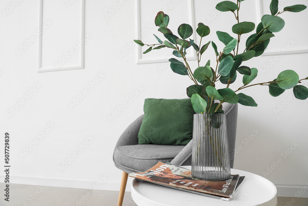 花瓶、杂志和扶手椅中有桉树的桌子，靠近浅色墙壁
