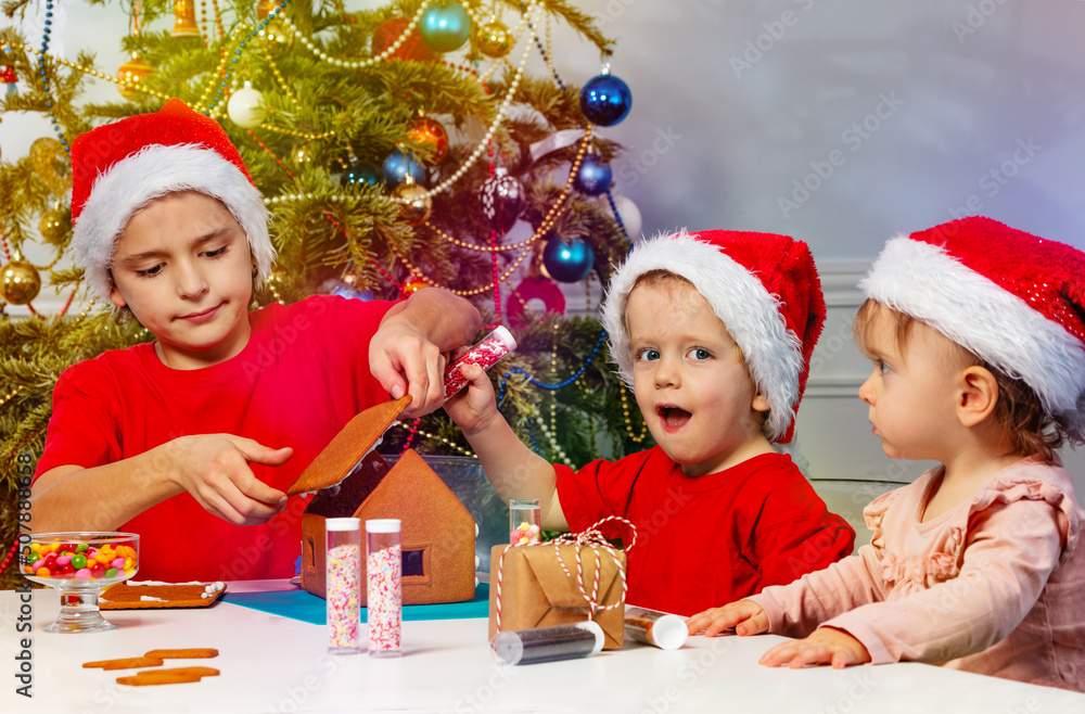 Two boys and girl decorate gingerbread house in Santa hats