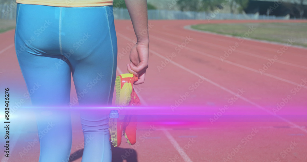 Image of light moving over midsection of woman holding running shoes walking on running track