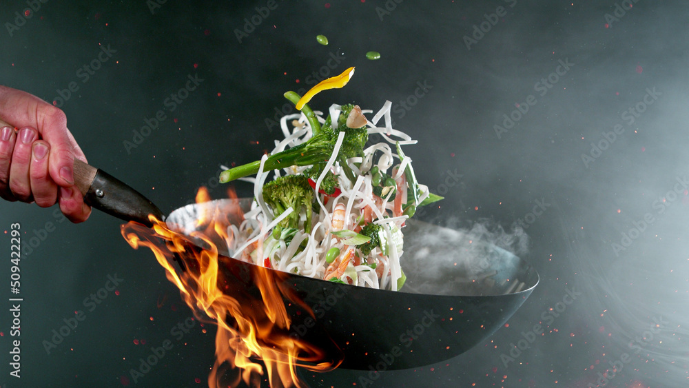 Closeup of chef throwing vegetable noodle mix from wok pan in fire.