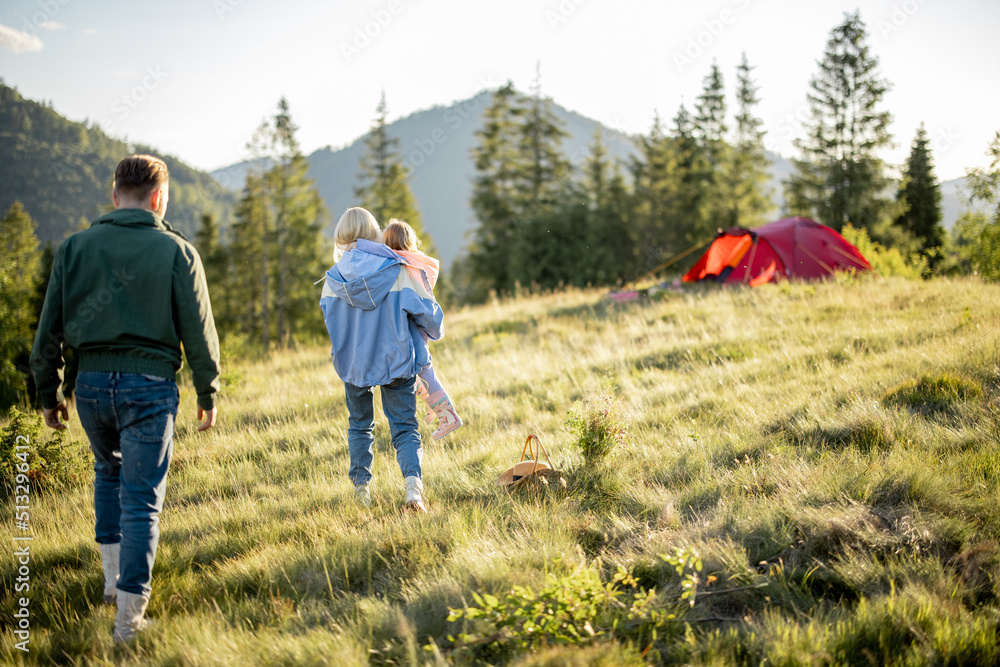 Young caucasian family with little girl walk together on green meadow while traveling with tent in t
