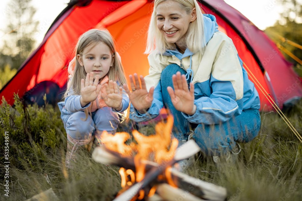 Young woman with her little girl sit by the campfire, bonding together while travel on nature. Mothe