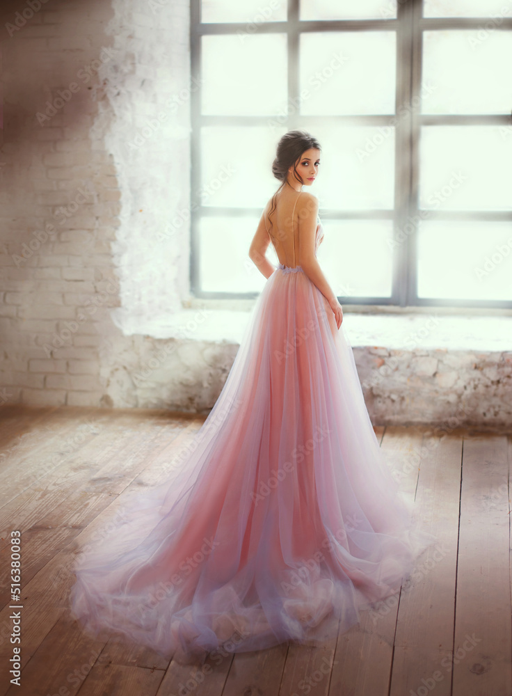 华丽的女孩时尚模特站在窗户附近，在阳光下等待爱情。傍晚的粉红色dre