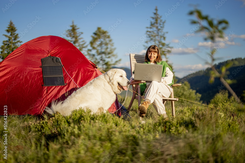 一名年轻女子带着帐篷和狗在山上旅行时用笔记本电脑工作。给电脑无线充电