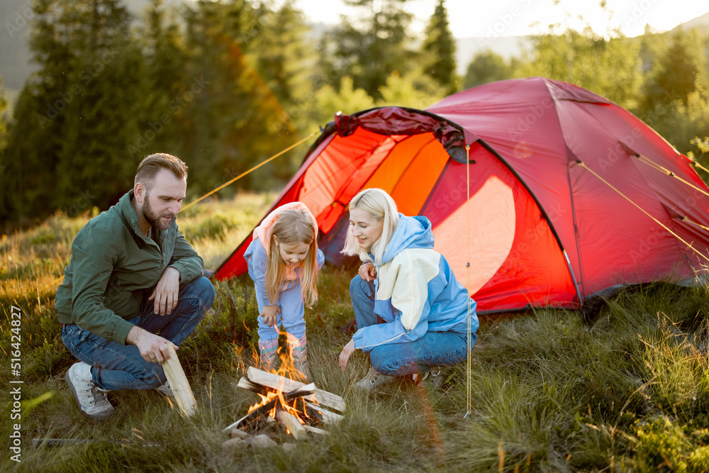 一对年轻的成年夫妇和一个小女孩坐在露营地的篝火旁，带着帐篷在大自然中旅行