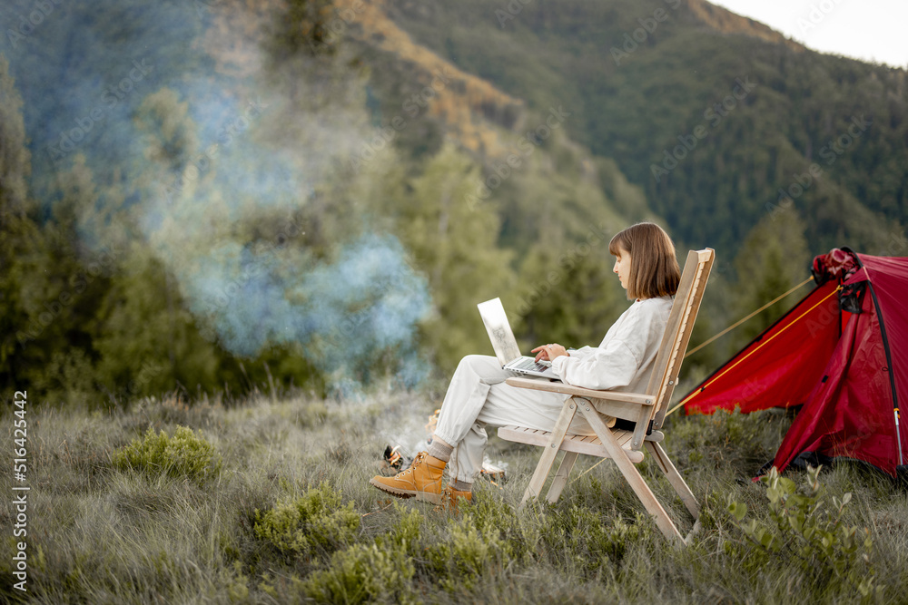 一名年轻女子在篝火旁的椅子上放松地坐着，带着帐篷旅行，一边用笔记本电脑工作