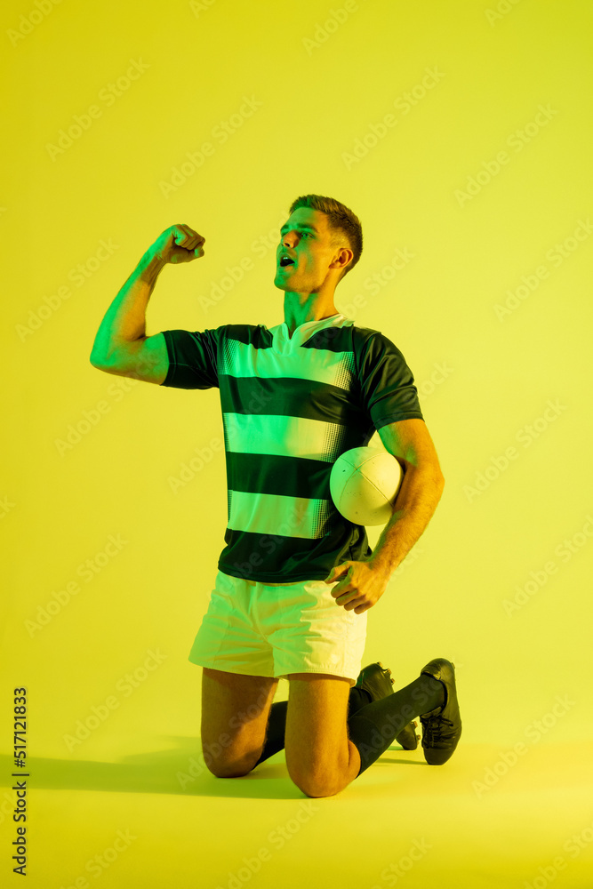 Happy caucasian male rugby player raising hands over yellow lighting