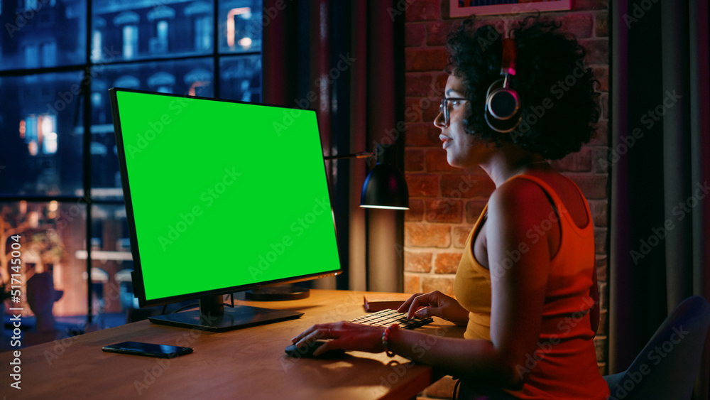 戴着耳机的年轻漂亮女性在家用绿色屏幕的台式电脑工作