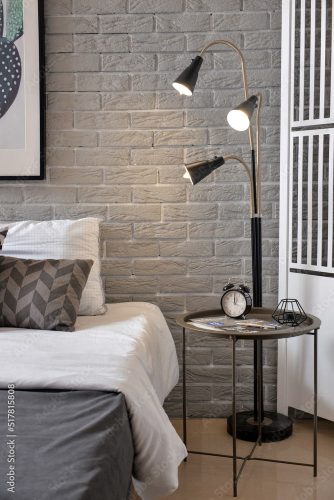 Table with alarm clock and glowing lamp near grey brick wall in bedroom