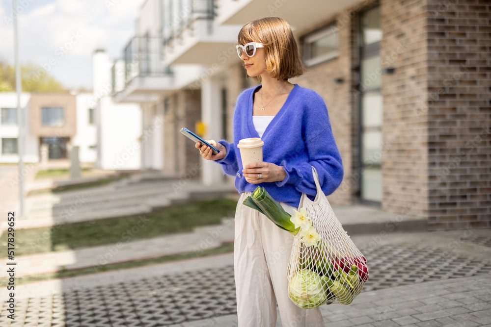 女人站着用手机，手里拿着装满新鲜蔬菜的网袋和可重复使用的咖啡杯