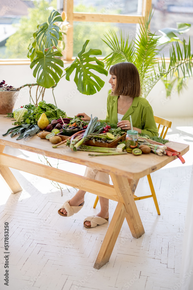 Portrait of a young cheerful woman sits by the table full of fresh vegetables, fruits and greens ind