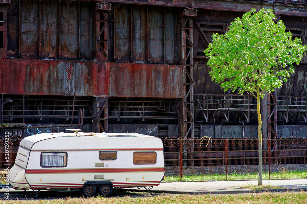 白色大篷车停在最后一棵年轻的绿树附近废弃钢铁厂锈迹斑斑的墙壁废墟上。查看t