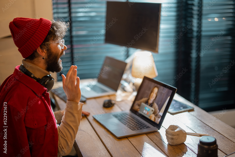 戴着红帽子的时尚男子坐在舒适的家庭办公室与女同事视频通话。Co