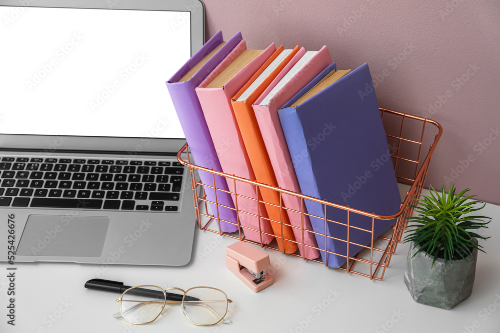 Holder with books, flowerpot, eyeglasses and laptop on table near pink wall
