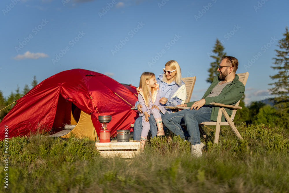 年轻的高加索夫妇带着小女孩放松地坐在椅子上，带着帐篷在山上旅行