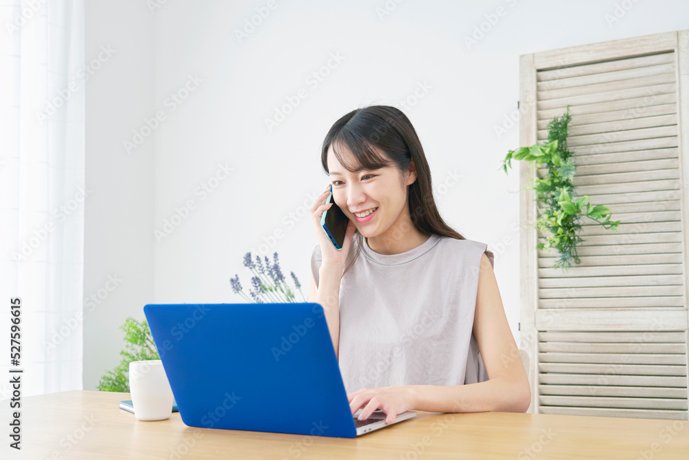 リビングでノートパソコンを見てスマホで電話する女性
