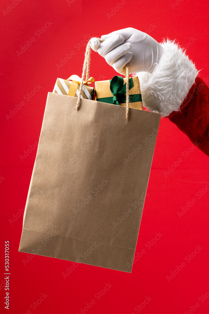 Image of hand of santa claus holding paper bag with christmas gifts on red background
