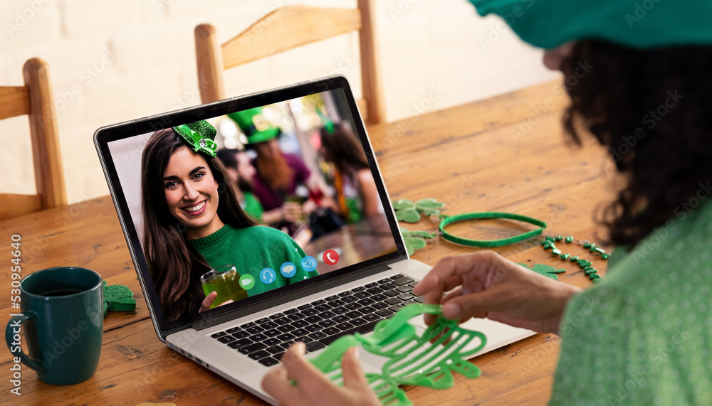 Caucasian woman holding shamrock glasses while having a video call on laptop at home
