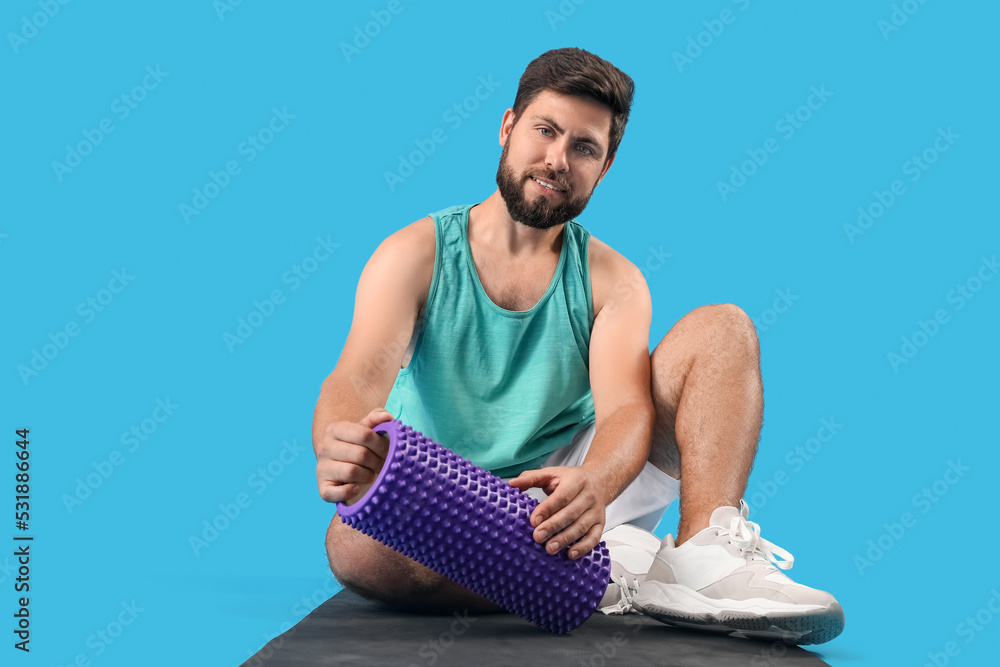 Young man with foam roller sitting on blue background