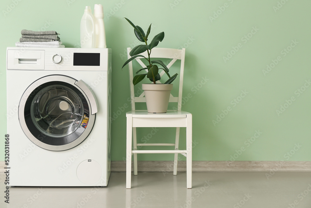 在靠近绿色墙壁的椅子上放有洗涤剂、毛巾和室内植物的洗衣机