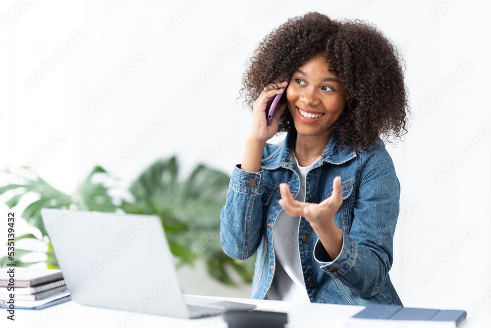 一位美丽的非洲妇女拿着笔记本电脑坐在办公室里，正在用手机聊天。Youn