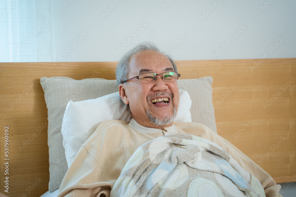 一位亚洲老人躺在家里卧室的床上，脸上露出开心的笑容。
