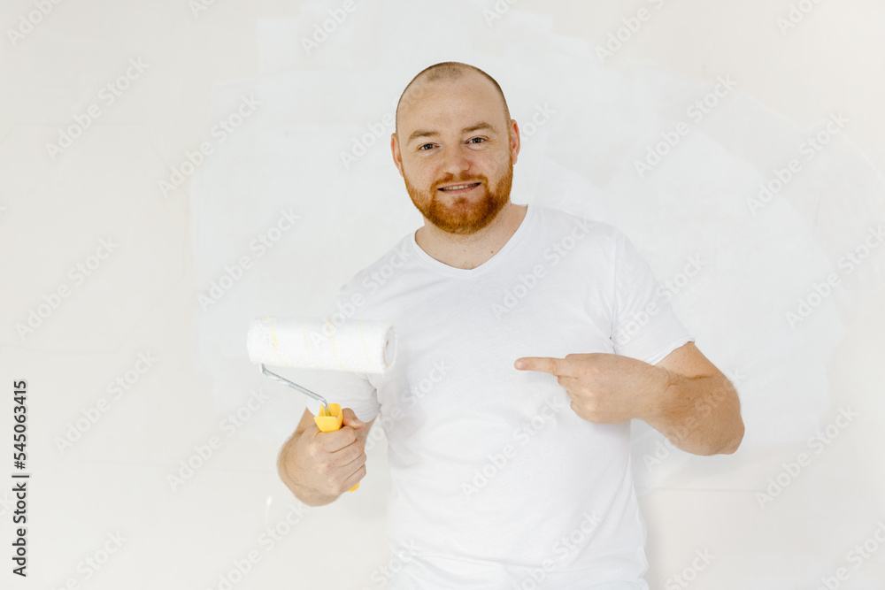 年轻有趣的微笑员工杂工男子穿着t恤，食指指向油漆滚筒。修理家