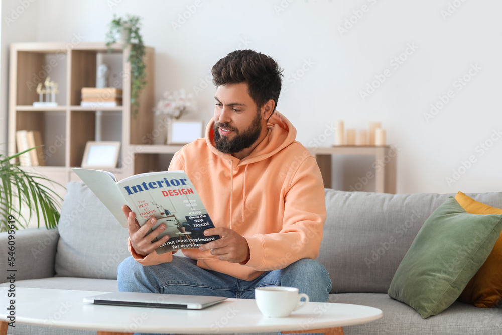 英俊的大胡子男人在家里的灰色沙发上看杂志